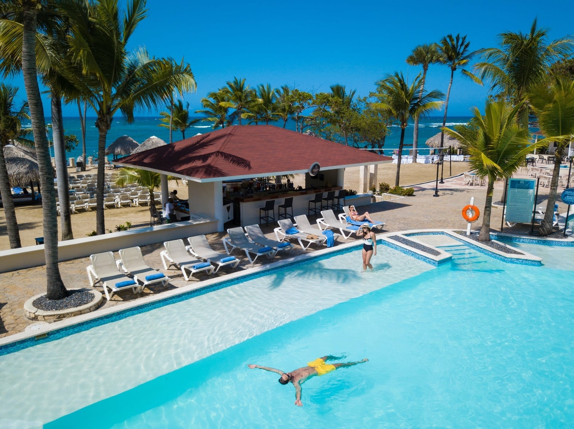 Aerial view of a tropical resort pool, bar, beach, and ocean with guests relaxing under palm trees.