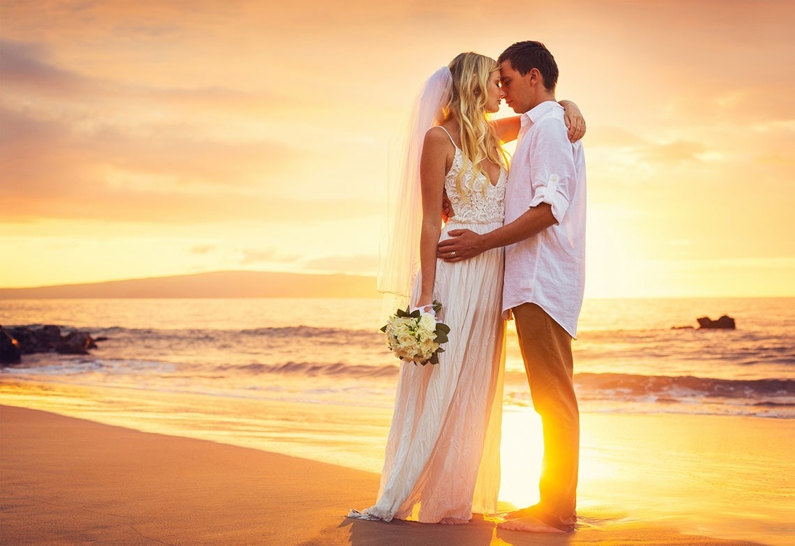 a bride and groom are kissing on the beach at sunset