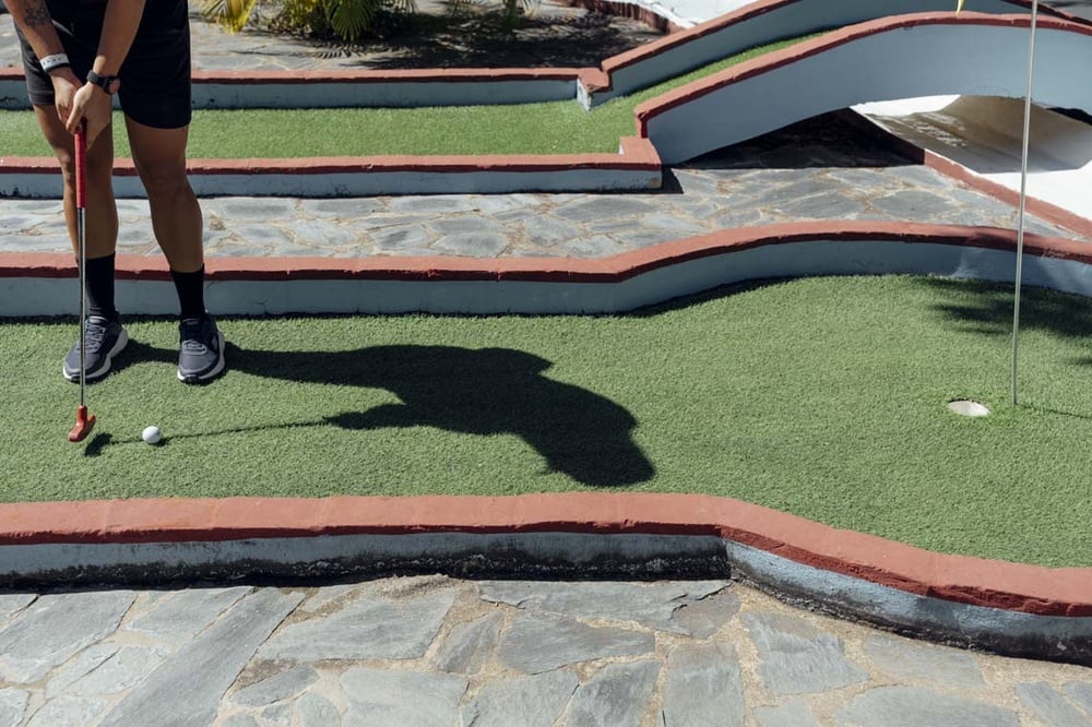 Minigolf player strikes ball at LHVC Resorts: Corpo, showing artificial turf, shadow, and bridge.