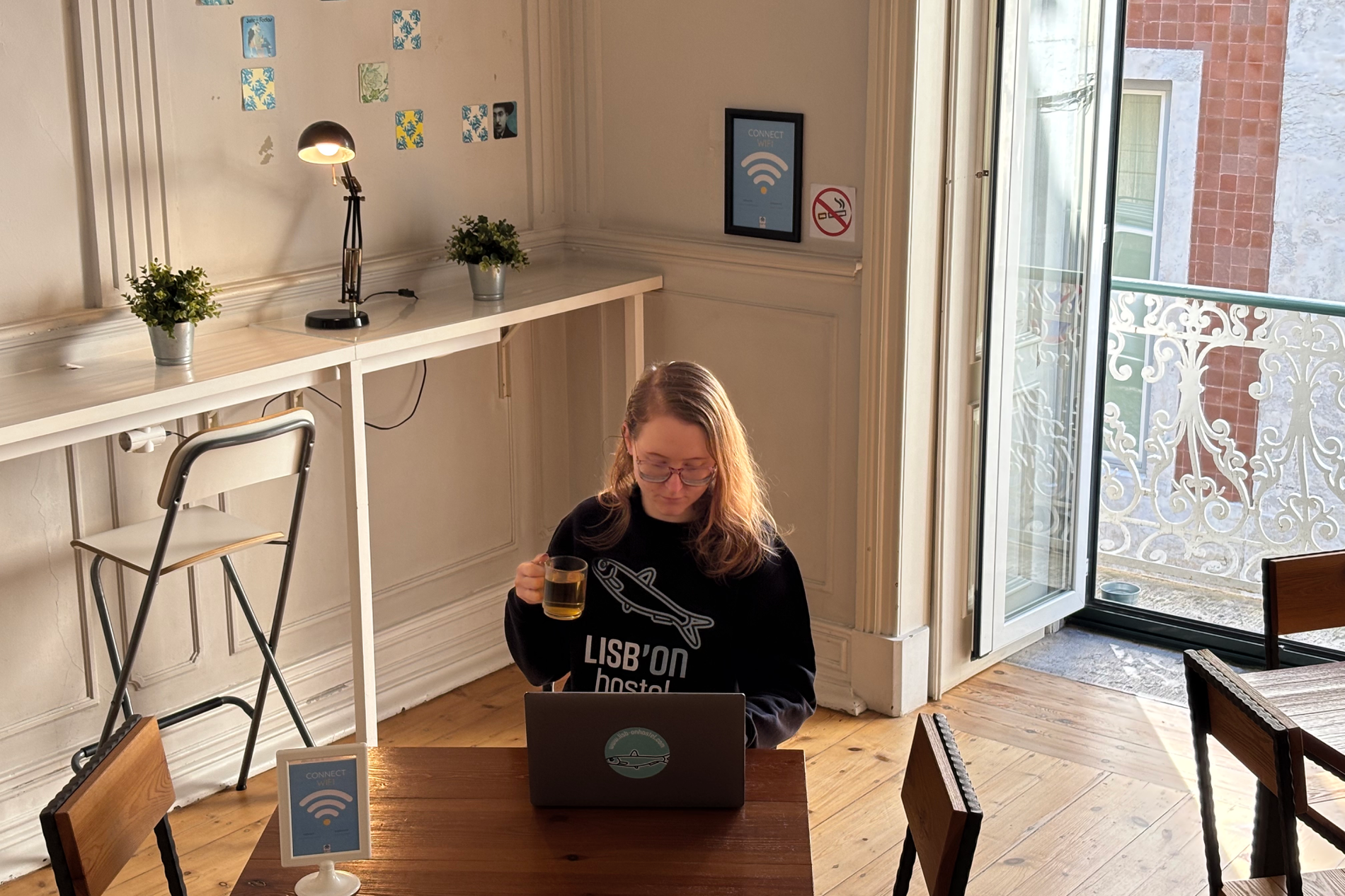 Una mujer con gafas y una sudadera de "LISB'ON hostel" trabaja en su portátil mientras bebe té, sentada en una mesa de madera en un luminoso espacio interior con un balcón al fondo.