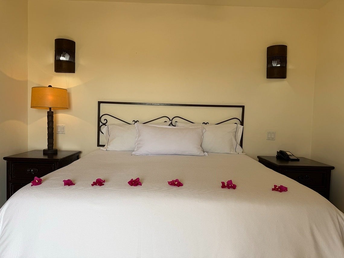 Una vista frontal de una cama de hotel hecha con sábanas blancas, flanqueada por mesitas de noche con lámparas y decorada con flores fucsias.