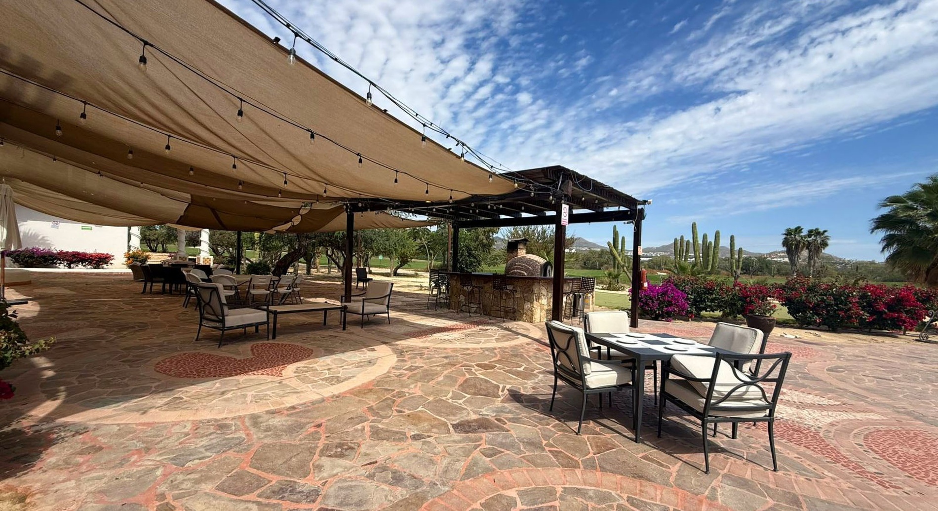 Una amplia terraza exterior sombreada con mesas, un bar con horno de pizza y vistas a un paisaje desértico de cactus y montañas.