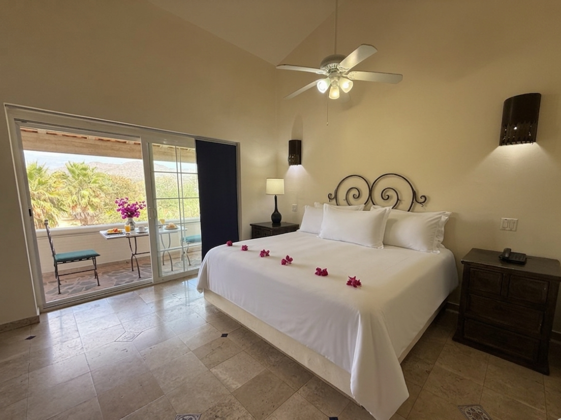 A spacious bedroom features a large bed with a wrought iron headboard, flanked by nightstands with lamps, a ceiling fan, and a tiled floor, with a sliding glass door opening to an outdoor area.