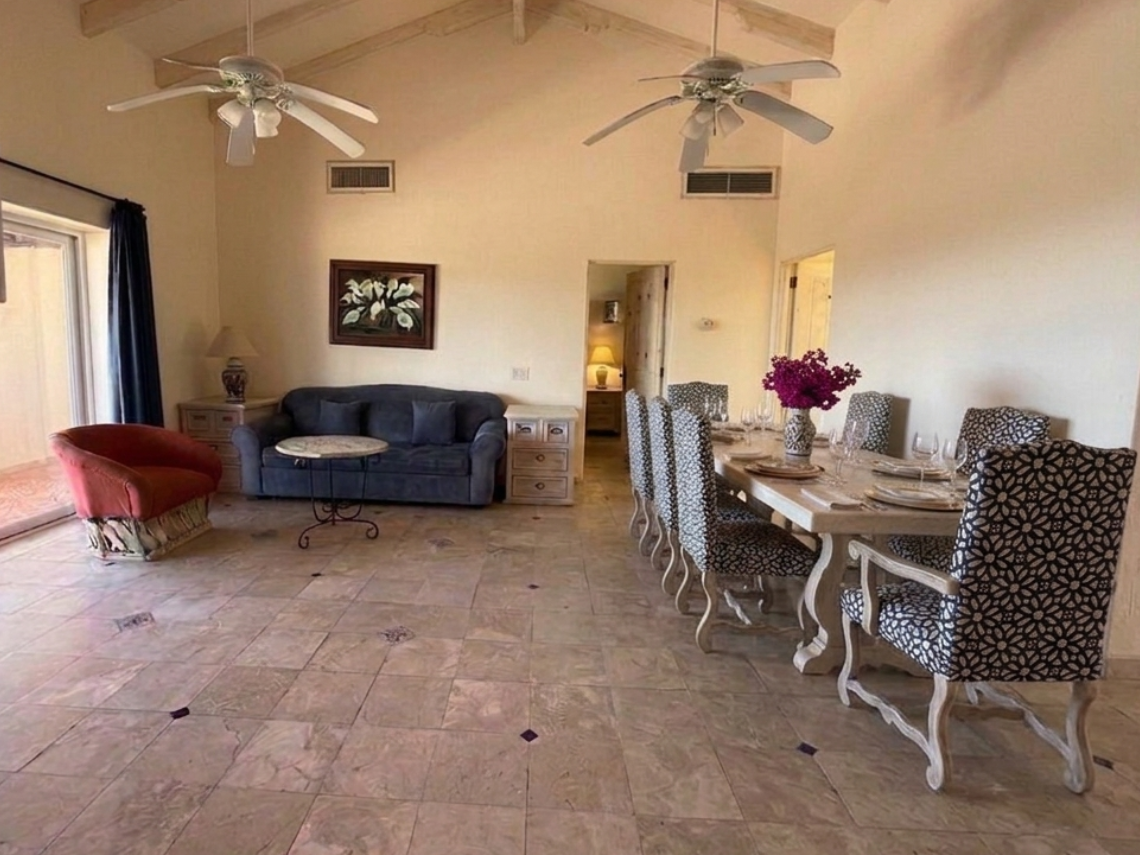 A spacious living and dining area with high beamed ceilings, tiled floors, and a mix of furniture, including a blue sofa, an orange armchair, and a dining table with patterned chairs, is captured in this bright interior shot.