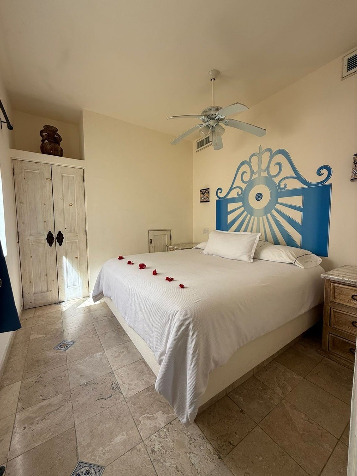 Un dormitorio luminoso con una cama grande adornada con pétalos de rosa, un cabecero azul pintado en la pared, un ventilador de techo y suelo de baldosas.