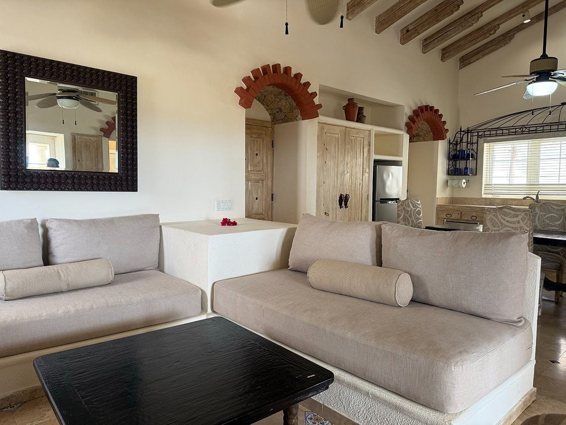 Un amplio salón-comedor presenta sofás de tonos neutros, una mesa de centro oscura y una decoración rústica mexicana con vigas de madera y detalles en ladrillo en las paredes.