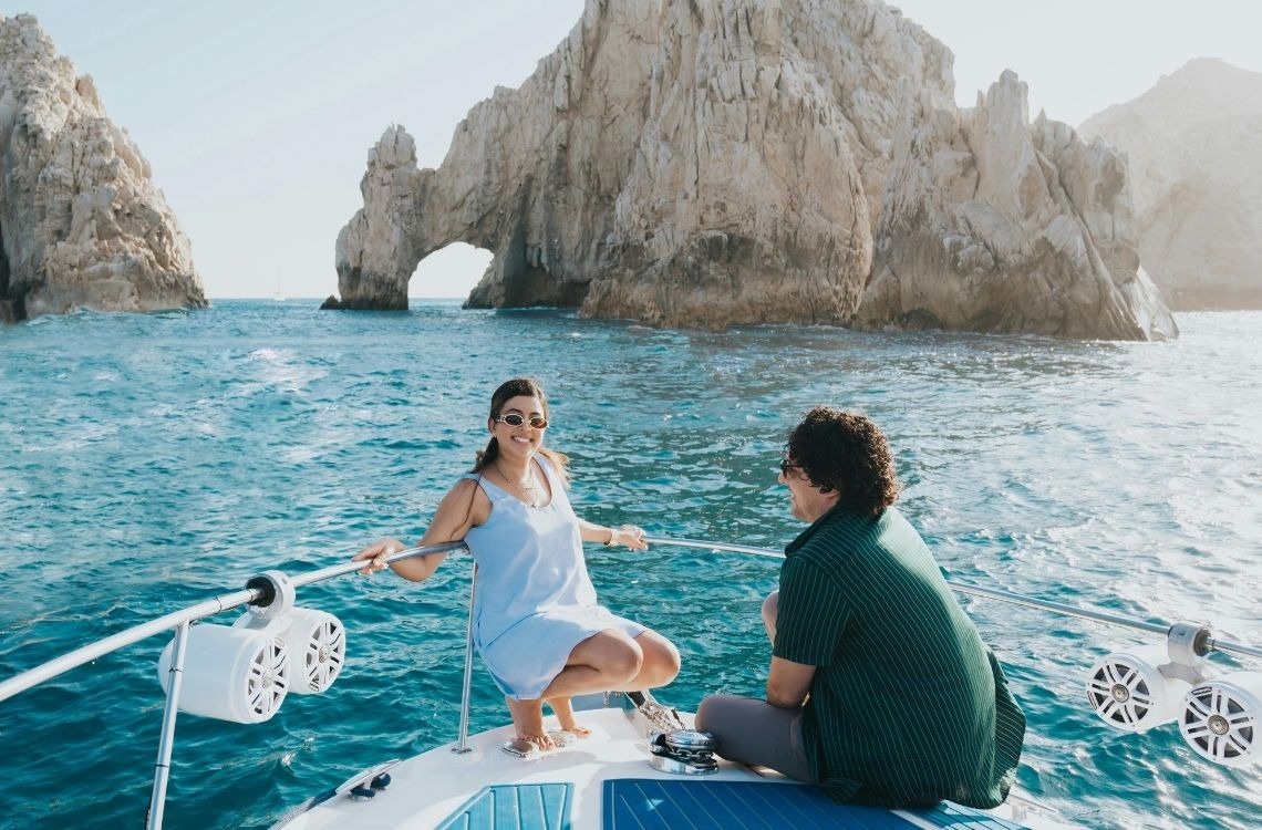 A resort pool area is lined with many white lounge chairs and umbrellas, set against a backdrop of Spanish-style buildings.