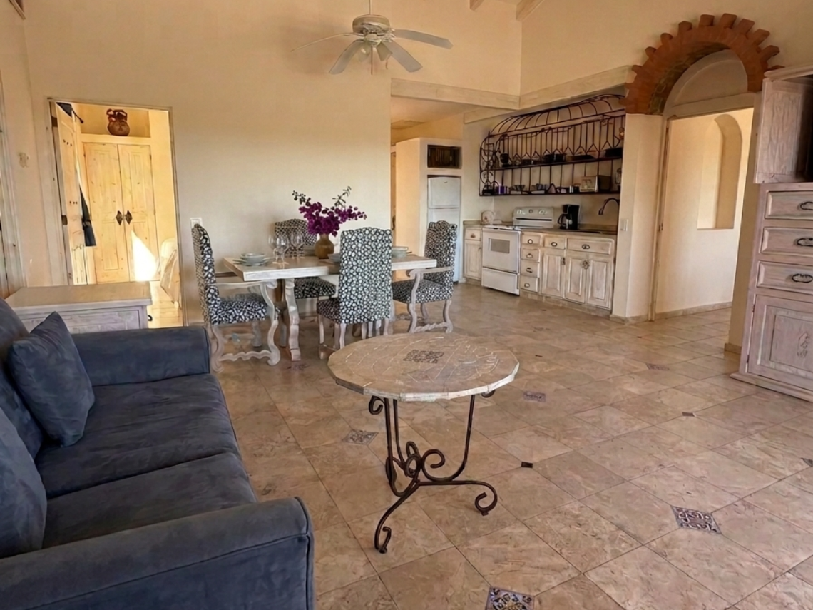 A bright and spacious open-concept living, dining, and kitchen area features vaulted ceilings with exposed beams, tiled floors, rustic-style furniture, and multiple arched doorways.