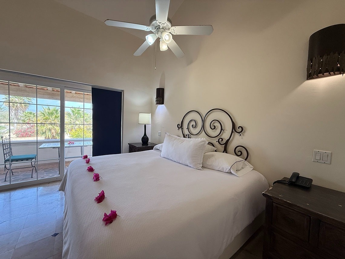 Un dormitorio luminoso con una cama grande adornada con flores rosas, un ventilador de techo y vistas a un patio exterior con abundante vegetación.