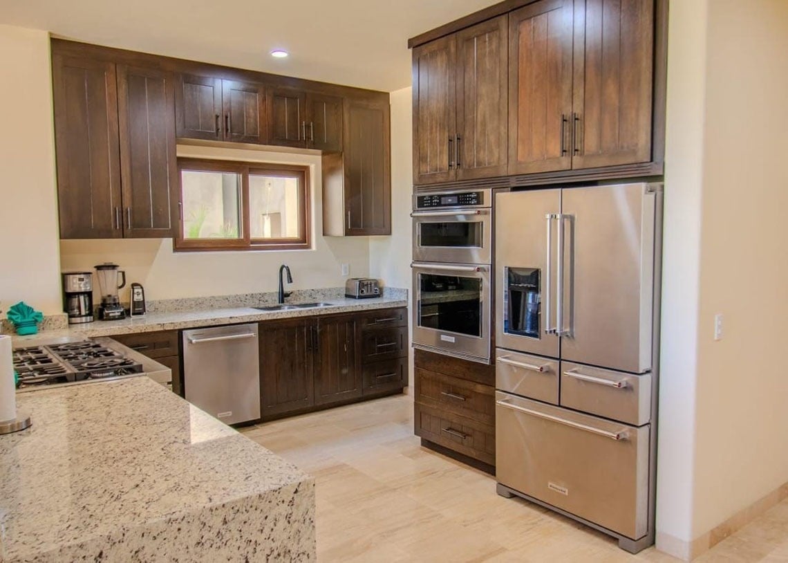 Cocina moderna con gabinetes de madera oscura, encimeras de granito claro y electrodomésticos de acero inoxidable, incluyendo una nevera, horno doble y lavavajillas.