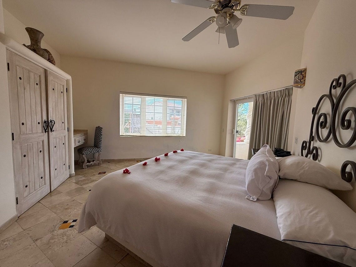 Se muestra una habitación espaciosa y luminosa con una cama adornada con pétalos de rosa, un armario de madera, una ventana con persianas y acceso a un patio exterior.