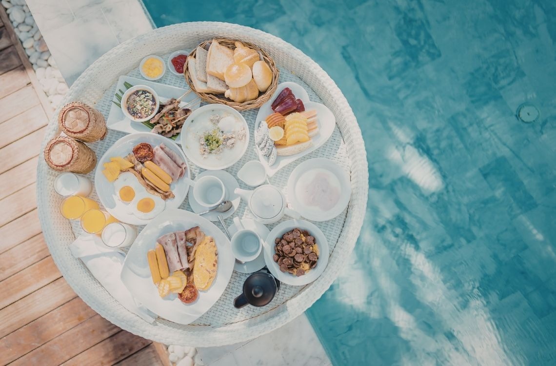 Una piscina de resort con tumbonas y sombrillas bajo el sol está rodeada de edificios de estilo mediterráneo o colonial.