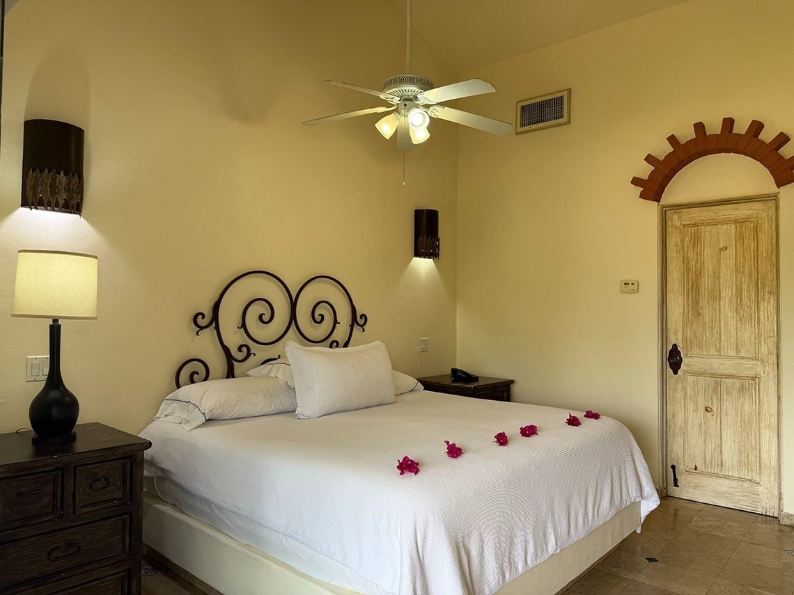 Una habitación luminosa presenta una cama grande con cabecero de hierro forjado y pétalos rojos, dos mesitas de noche con lámparas, un ventilador de techo y una puerta de madera rústica con un arco decorativo.