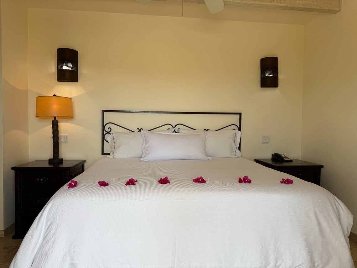 Una cama doble con sábanas blancas está adornada con pétalos de flores y flanqueada por mesitas de noche con lámparas y apliques en la pared.