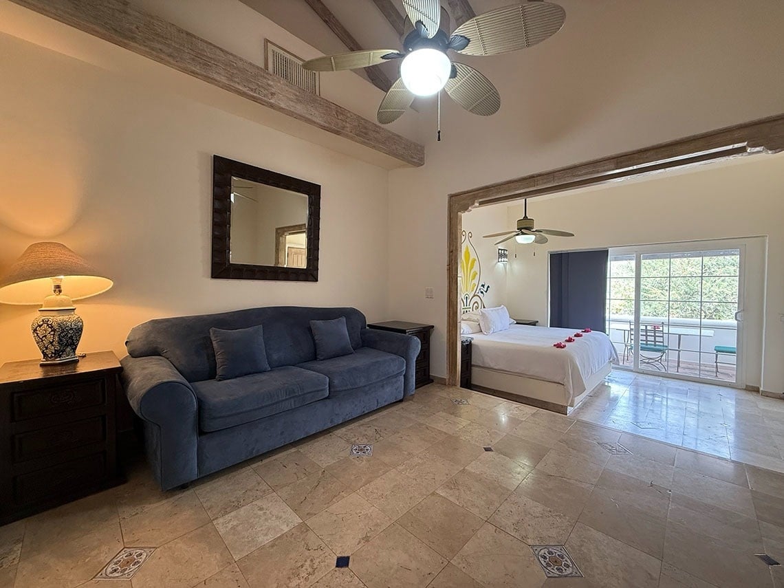 Una suite de hotel luminosa con una sala de estar que incluye un sofá azul y un dormitorio con cama blanca y balcón, separados por un arco, con suelos de baldosas y ventiladores de techo.