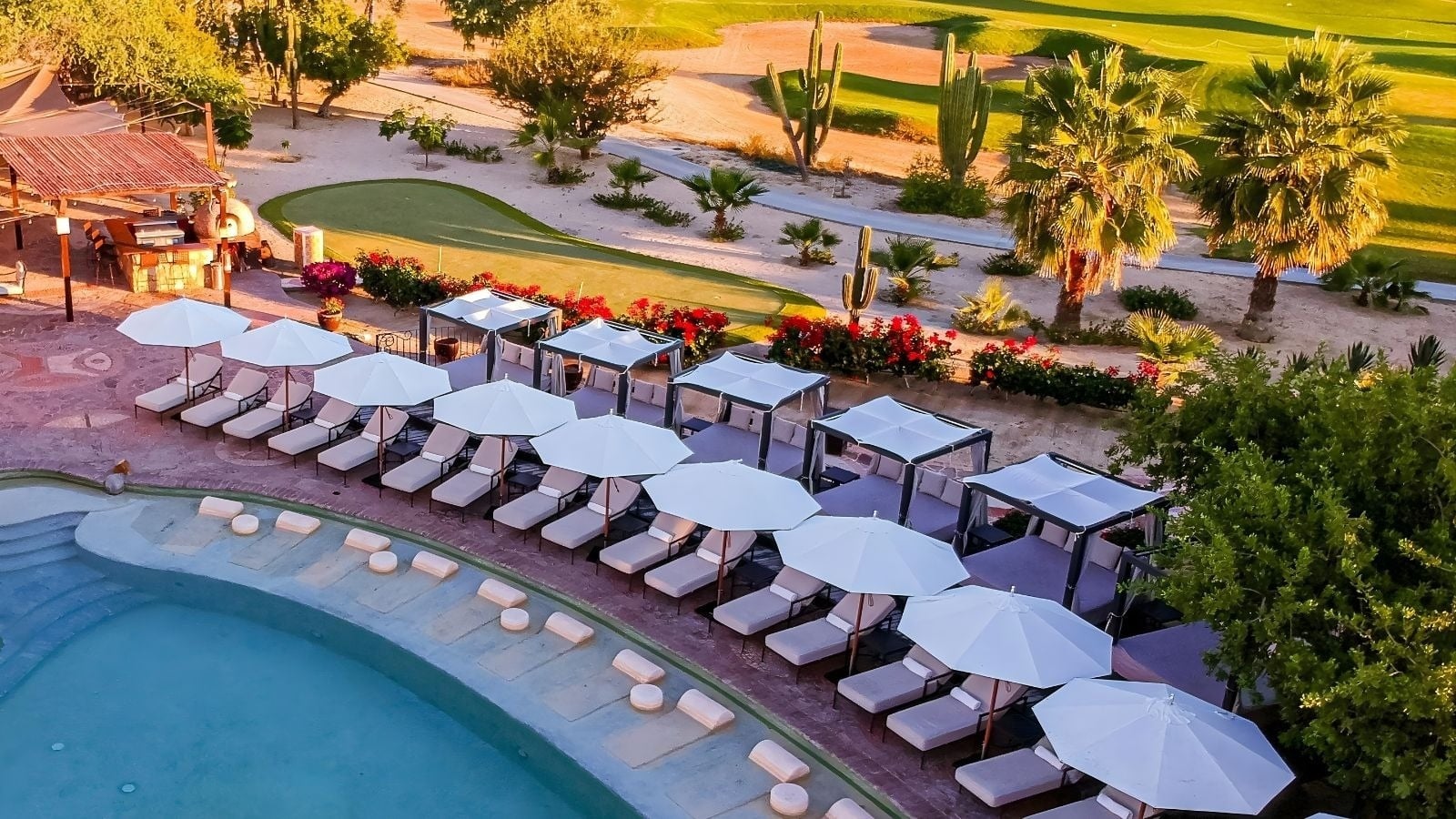 Vista aérea de un lujoso resort con piscina, tumbonas con sombrillas y cabañas junto a un campo de golf árido salpicado de cactus y palmeras, todo bañado por la cálida luz del atardecer.