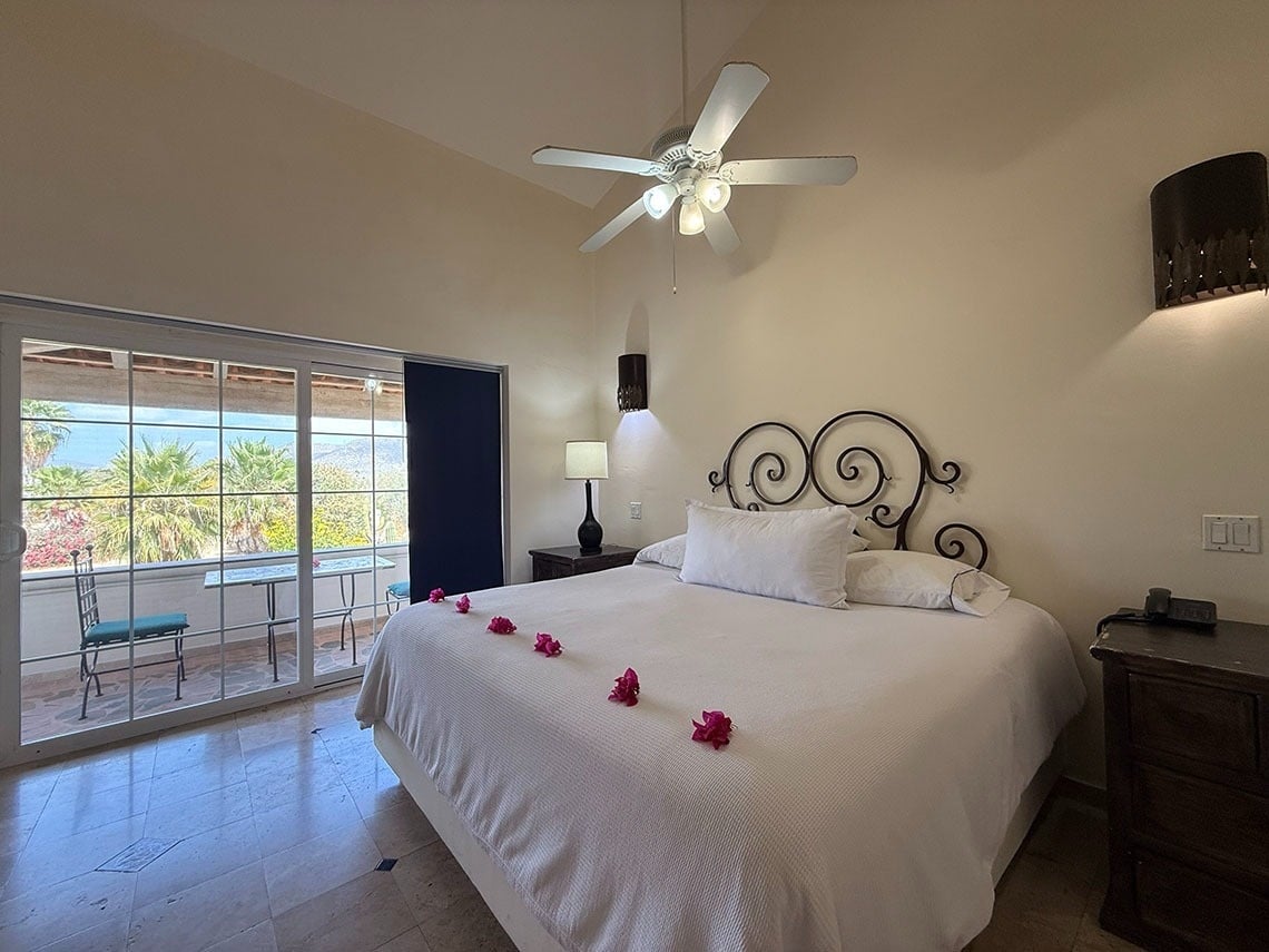 Un dormitorio luminoso con cama de hierro forjado, ventilador de techo y balcón con vistas tropicales a palmeras y montañas.
