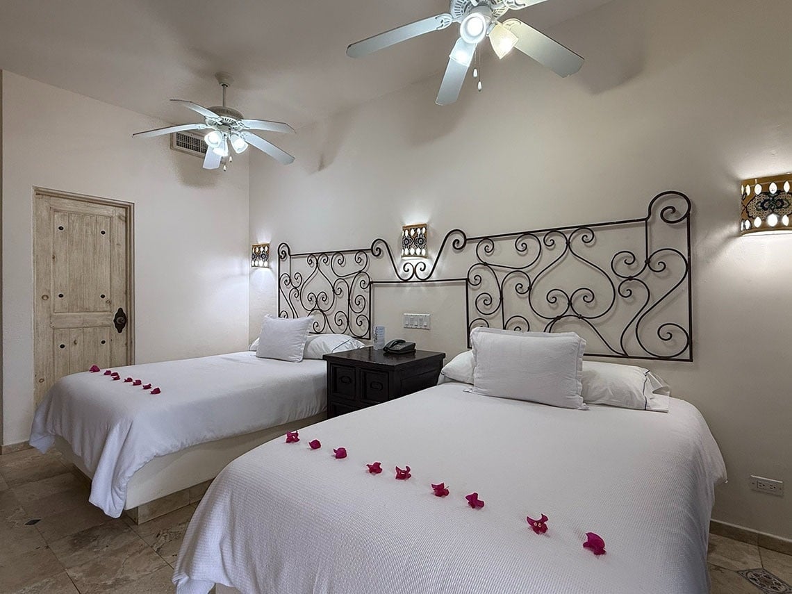 Una habitación de hotel con dos camas individuales decoradas con pétalos, cabeceros de hierro forjado, ventiladores de techo y una puerta de madera rústica.