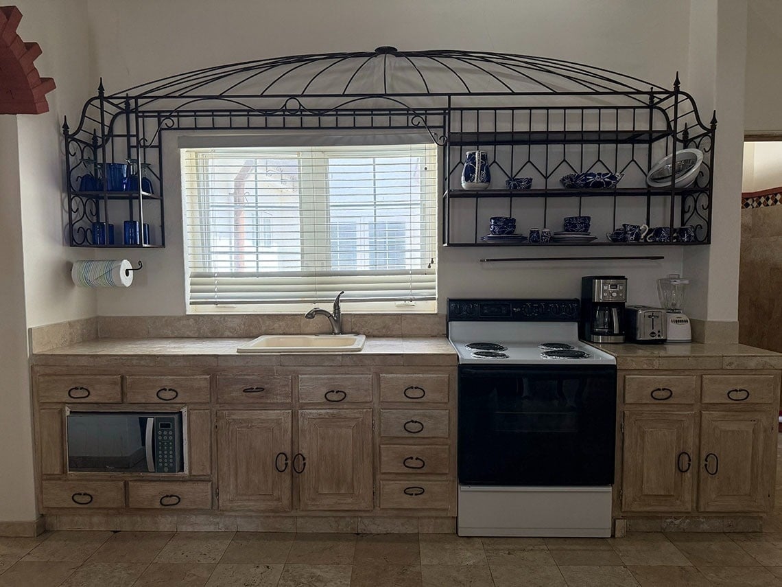 Una cocina rústica con gabinetes de madera clara, encimeras de azulejos, un fregadero, una estufa, un microondas empotrado y estantes de metal forjado decorados con vajilla.