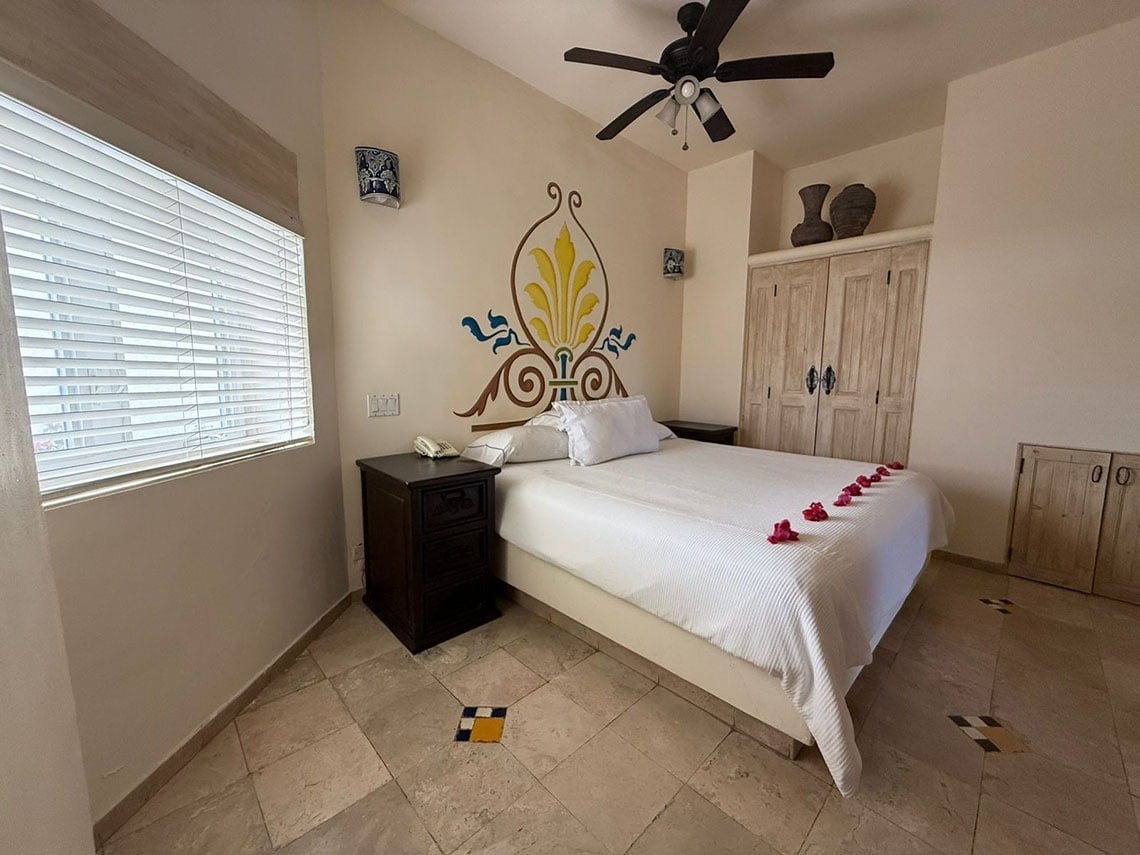 Una habitación con una cama blanca adornada con pétalos de rosa, un cabecero decorado, persianas en la ventana y un suelo de baldosas con incrustaciones.