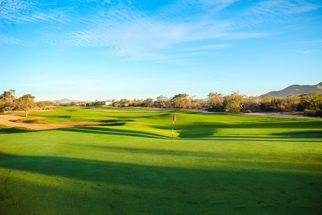 Una vista panorámica de un exuberante campo de golf con césped verde vibrante, largas sombras, una bandera roja y un cielo azul claro, flanqueado por árboles y montañas distantes bajo la luz del sol.