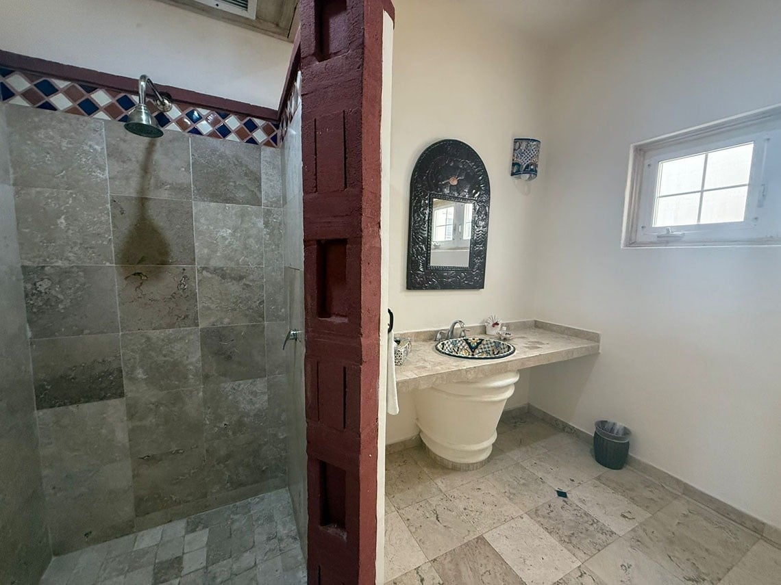 Un cuarto de baño con una ducha de azulejos, un tocador de piedra con lavabo decorativo y un espejo, separado por una pared de ladrillo pintada de rojo.