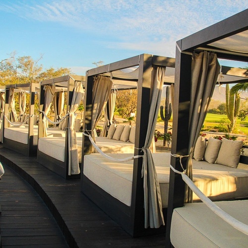 Una hilera de camas balinesas con cortinas se alinea en una terraza de madera, ofreciendo vistas a un campo de golf con cactus bajo un cielo soleado.