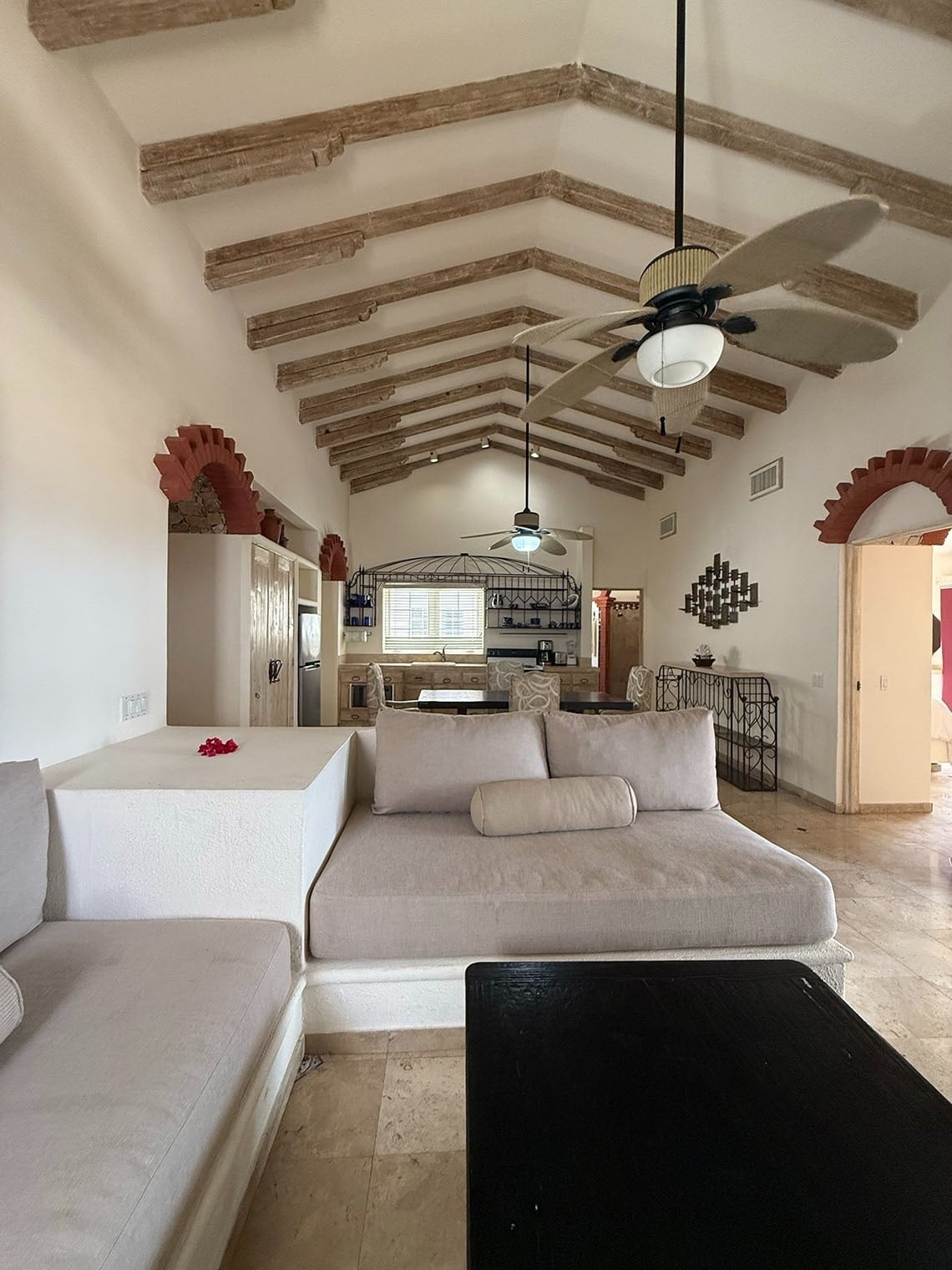 Una sala de estar de estilo rústico presenta sofás empotrados, un techo abovedado con vigas de madera expuestas y ventiladores de techo, con detalles de arcos de ladrillo y un área de cocina/comedor visible al fondo.