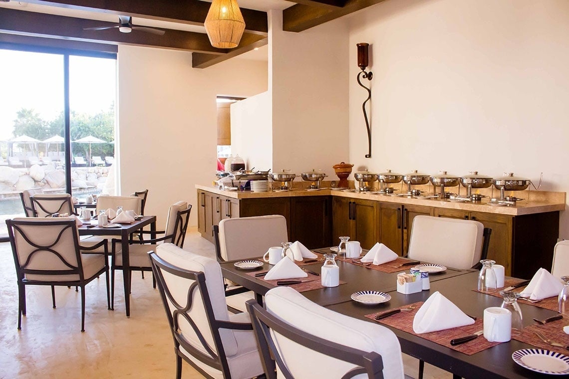 Un comedor luminoso cuenta con mesas puestas, un buffet y grandes ventanales con vistas a una piscina exterior.