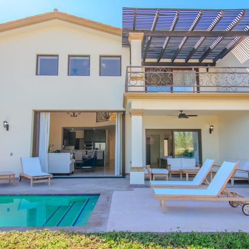 An aerial view captures a modern white house with a swimming pool and lush green lawn, set against a backdrop of a dry, arid landscape with sparse vegetation and distant hills.