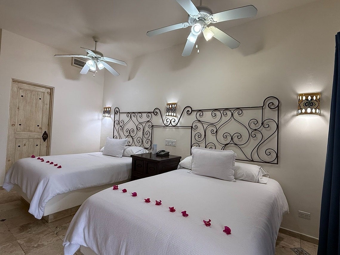 Una habitación de hotel con dos camas individuales decoradas con pétalos de flores rojas, cabeceros de hierro forjado ornamentados, ventiladores de techo y luces de pared decorativas.