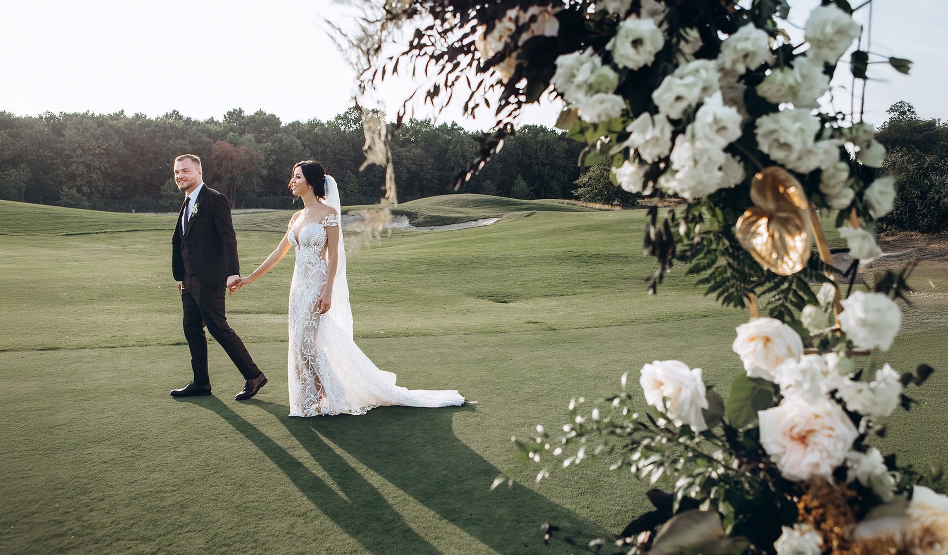 Una pareja de recién casados camina de la mano por un campo de golf al atardecer, con una decoración floral en primer plano a la derecha.