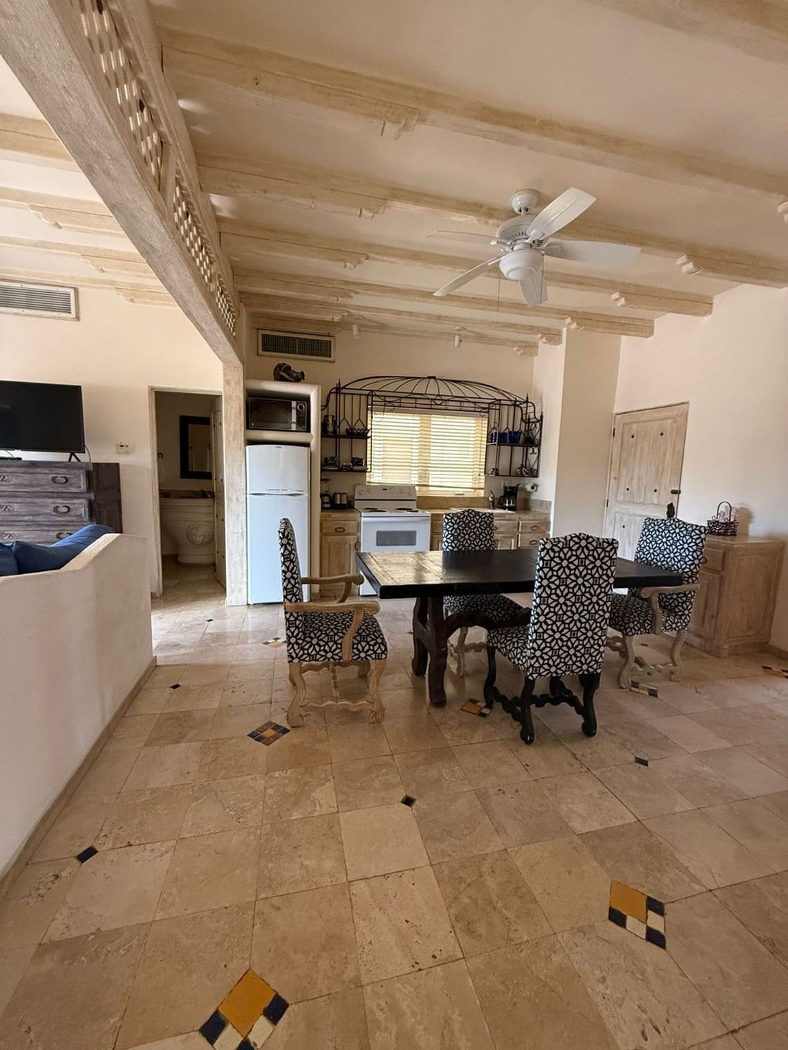 Un espacio abierto combina sala de estar, comedor y cocina, con vigas de madera expuestas en el techo, un ventilador y suelo de baldosas decoradas.