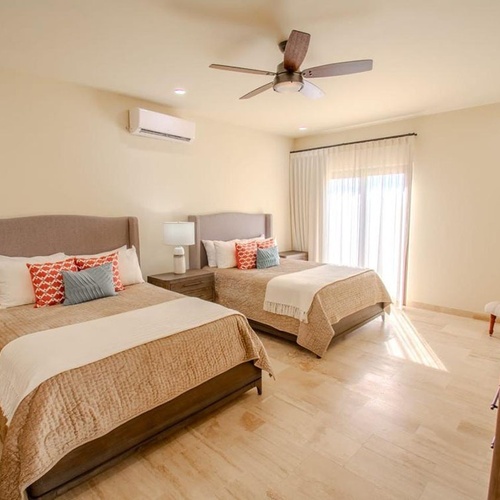 Una habitación luminosa con dos camas individuales, ventilador de techo, aire acondicionado y grandes ventanales con cortinas transparentes.