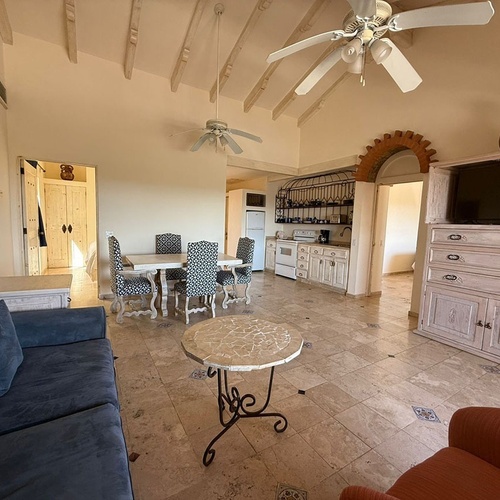 Una amplia sala de estar y comedor de estilo rústico-mediterráneo presenta techos abovedados con vigas, ventiladores de techo, mobiliario de madera clara y un sofá azul, con una cocina visible al fondo a través de un arco de ladrillo.