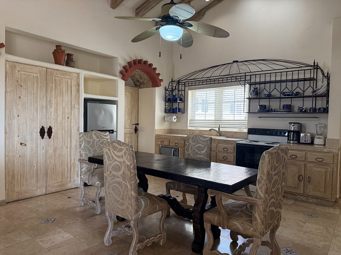 Una cocina-comedor de estilo rústico-mediterráneo con un gran ventilador de techo, mesa de comedor con sillas tapizadas, gabinetes de madera clara, electrodomésticos negros y plateados, y estanterías decorativas de hierro forjado sobre la ventana.