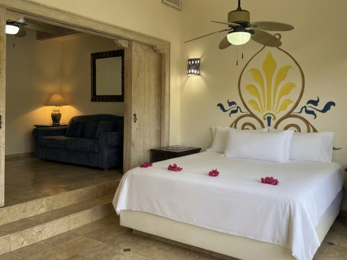A bright bedroom with a bed adorned with red petals and a decorative wall mural is shown, with a ceiling fan overhead and an adjacent sitting area with a sofa visible through an open doorway and steps.