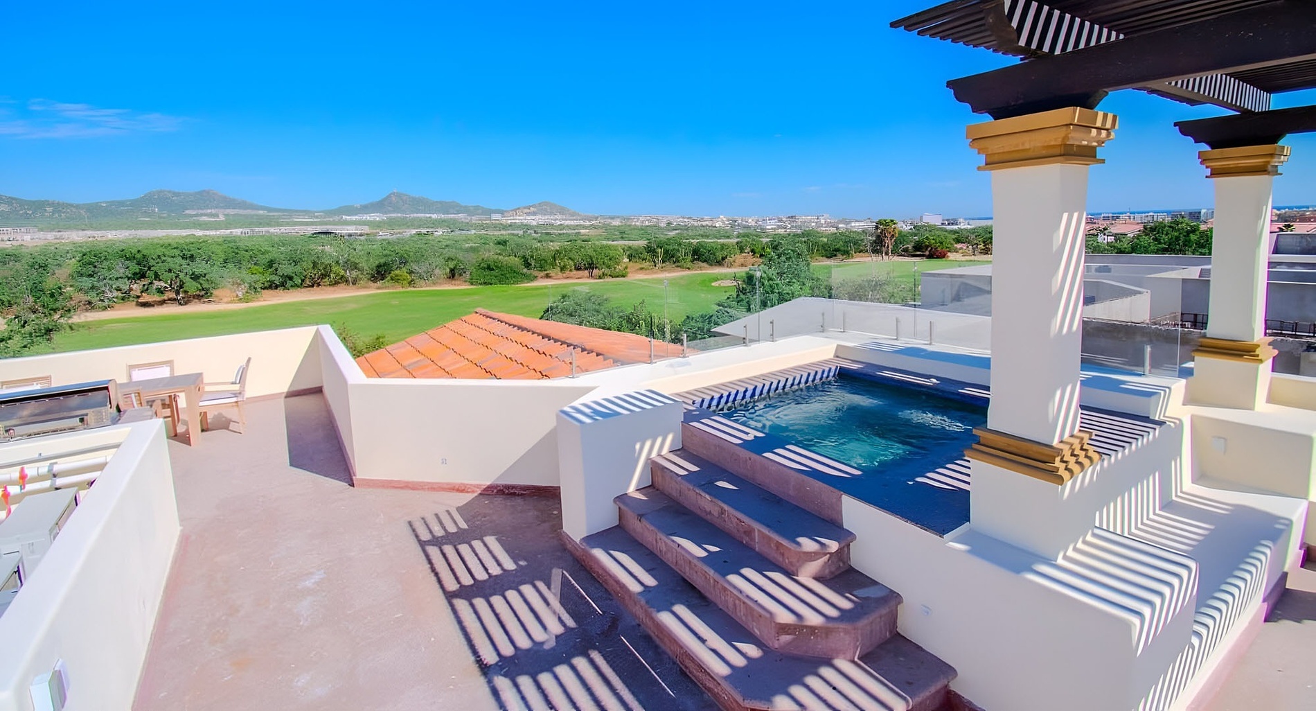 Una terraza en la azotea con una pequeña piscina, comedor al aire libre y parrilla ofrece vistas panorámicas a un campo de golf y montañas bajo un cielo azul claro.
