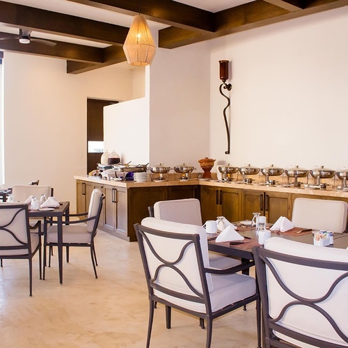 Una vista de un comedor interior con mesas y sillas puestas, un bufet con calentadores de comida y grandes ventanales con vista a una zona de piscina exterior.