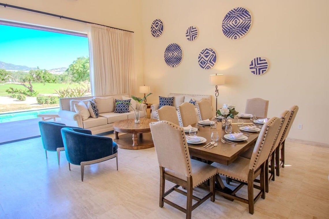 Interior moderno de sala de estar y comedor abierto con ventanales a una piscina y jardín, decorado con muebles claros y detalles en azul.