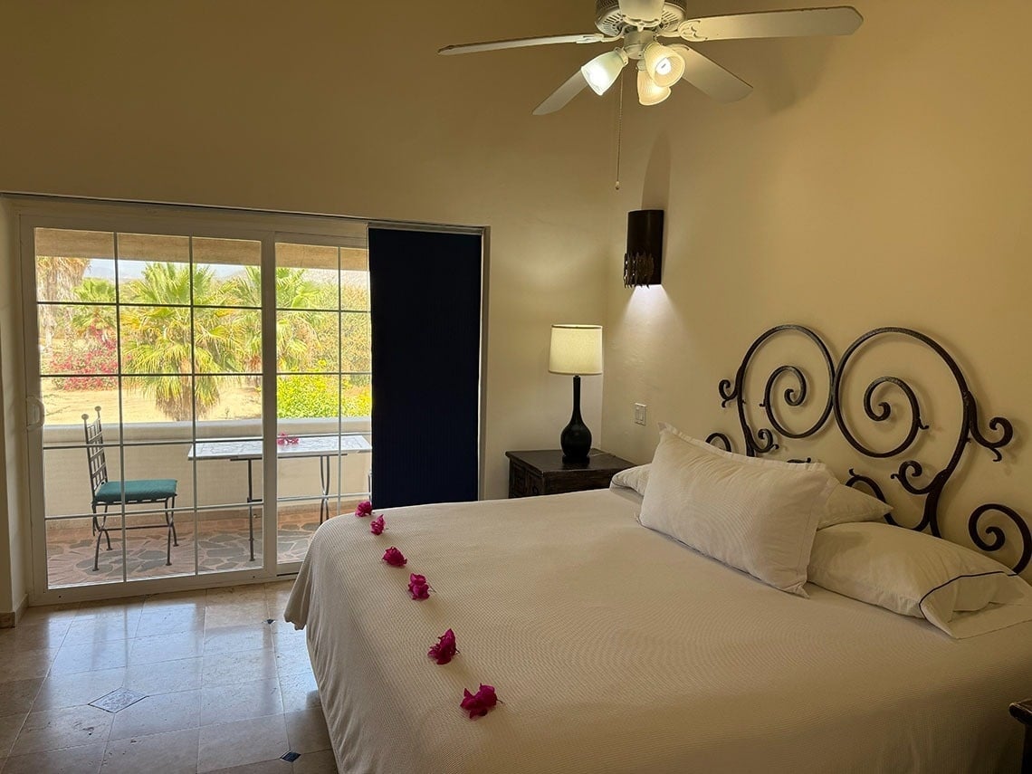 Habitación con una cama decorada con pétalos de flores, con acceso a un balcón amueblado que ofrece vistas a palmeras y exuberante vegetación.