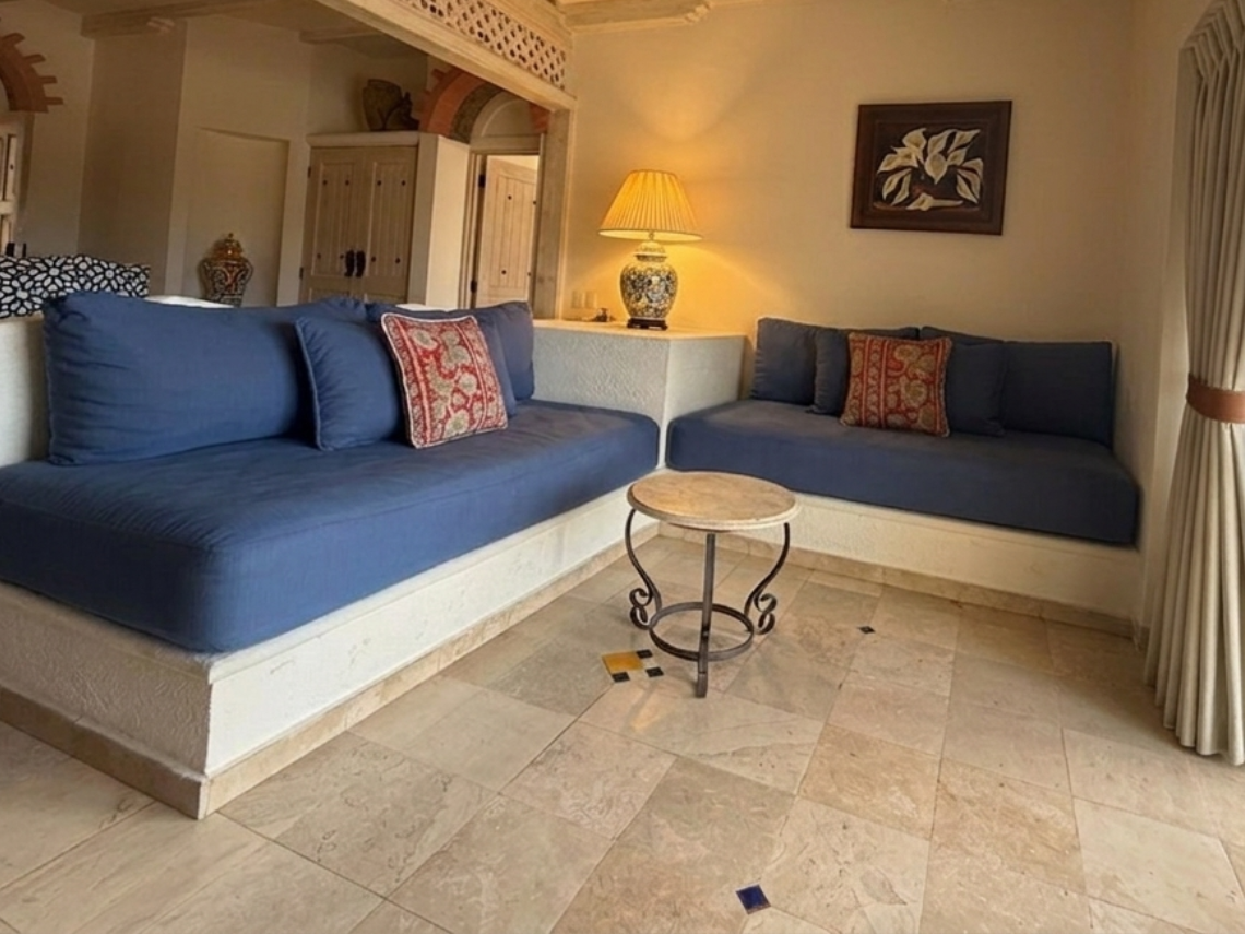 An L-shaped living area features built-in blue sofas, light-colored tiled flooring, and a ceiling with exposed wooden beams and lattice work, with a sliding glass door opening to an outdoor patio.