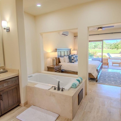 Un amplio baño con bañera integrada y tocador se conecta a un luminoso dormitorio que ofrece vistas a un jardín exterior.