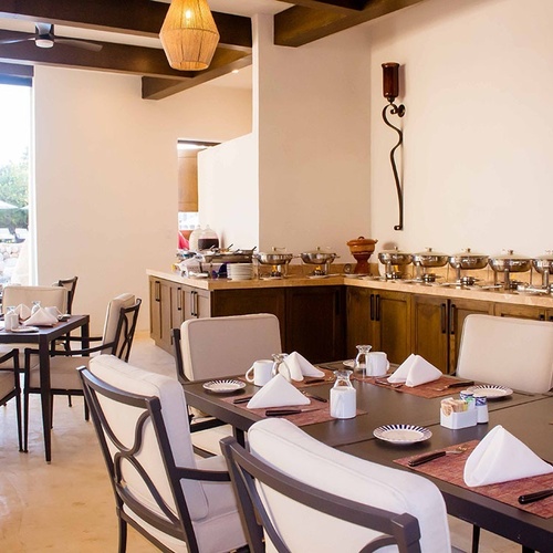 Una sala de comedor interior con un bufé de comida y mesas puestas, con grandes ventanales que dan a una zona de piscina exterior.