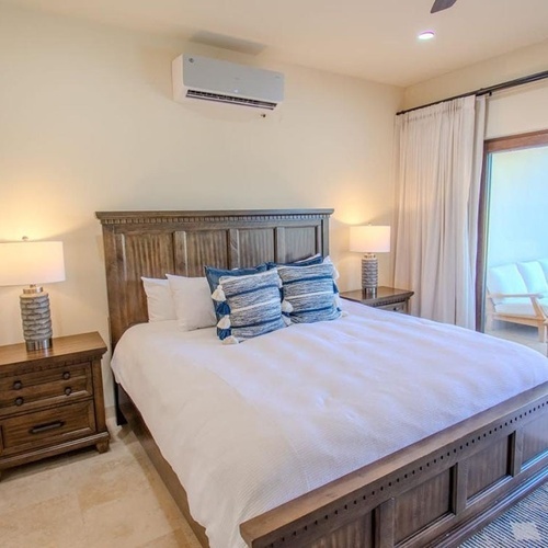 Dormitorio luminoso con una cama de madera grande, mesitas de noche con lámparas, aire acondicionado y acceso a un patio exterior amueblado a través de una puerta corredera de cristal.
