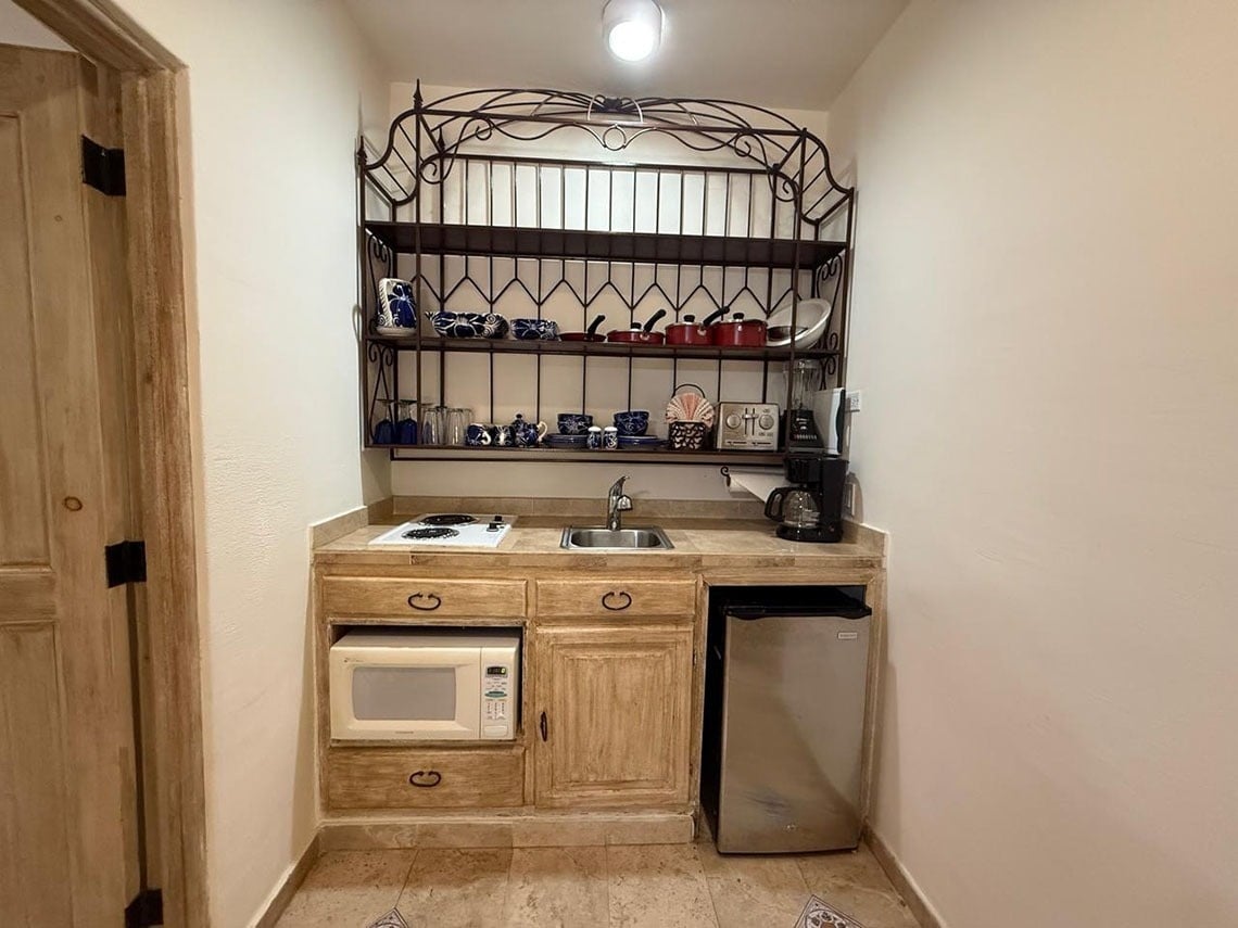 Una pequeña cocina de estilo rústico presenta gabinetes de madera, una encimera con fregadero y estufa, un microondas empotrado, una mini-nevera y una estantería de hierro forjado llena de utensilios.