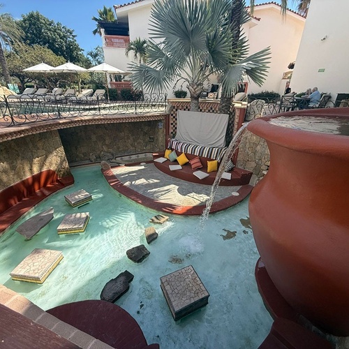 La imagen muestra un área de resort al aire libre con una gran fuente de agua que cae en un canal con escalones, una zona de asientos hundida y una piscina con tumbonas en segundo plano.