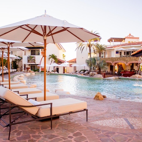 Una lujosa piscina de resort con tumbonas bajo sombrillas, edificios de estilo mediterráneo y un bar acuático con cascada.