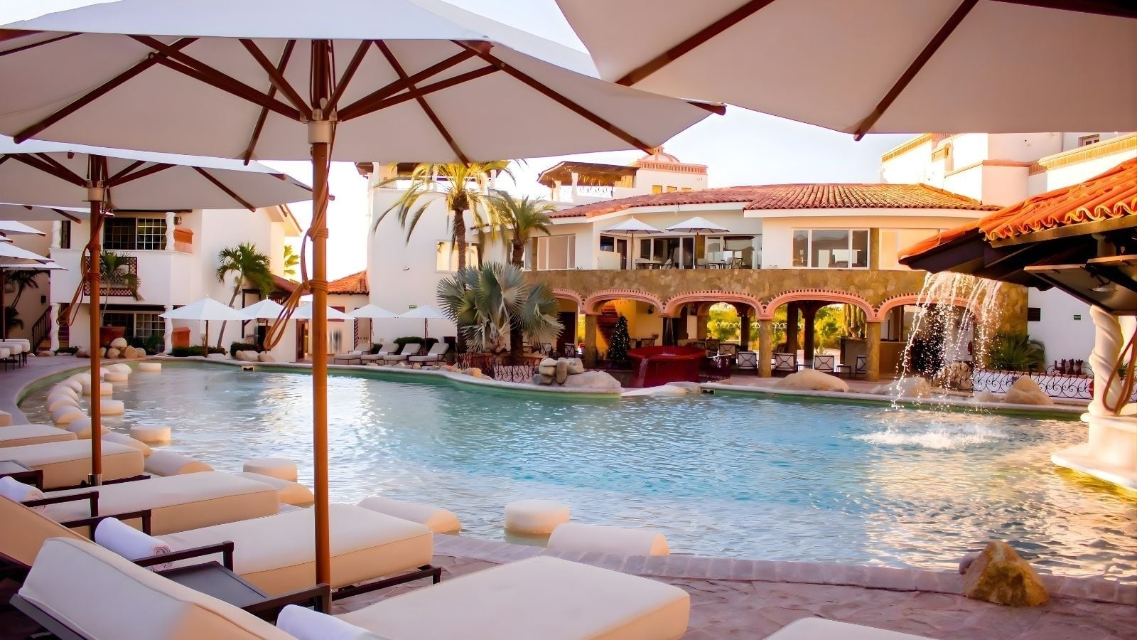 Una vista de una lujosa zona de piscina de un resort con tumbonas, sombrillas, edificios de estilo mediterráneo con tejados rojos, palmeras y una fuente de agua bajo la luz del sol.
