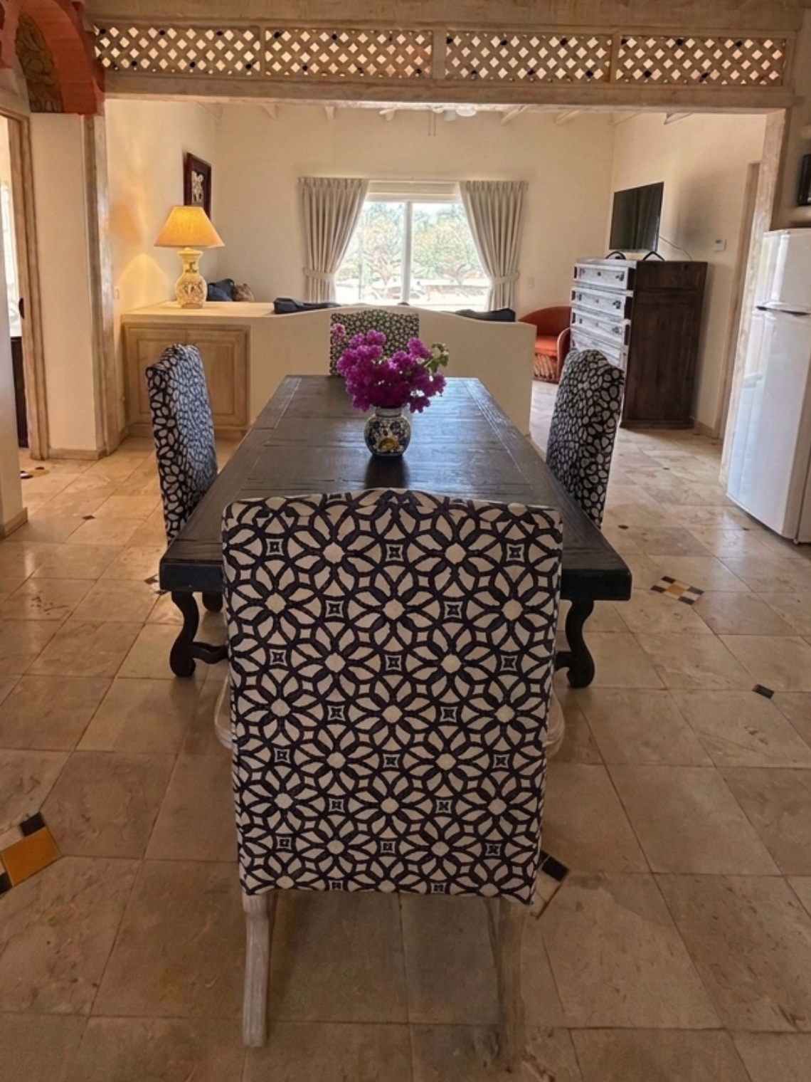 An open-concept living space features an earthy-toned tiled floor with decorative accents, exposed wooden ceiling beams, a dining area with patterned chairs, and a visible kitchenette.